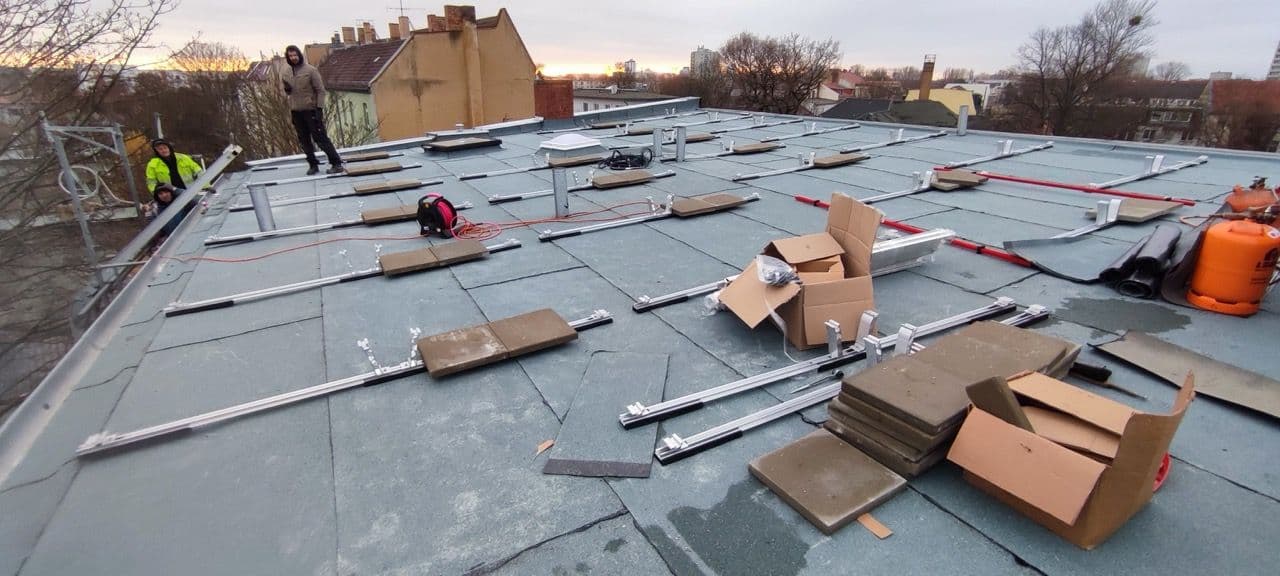 Auf einem Dach in Berlin werden Schienen verlegt, um die PV Anlage zu montieren. Zwei Mitarbeiter der Firma sunday solar sind zu sehen. Im Hintergrund rosa Wolken und Bäume ohne Laub